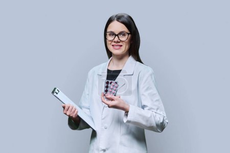 Photo for Young woman doctor in white coat with clipboard showing medicine pills in blisters looking at camera on gray studio background. Medicine, treatment, pharmacology, pharmaceuticals, health care concept - Royalty Free Image