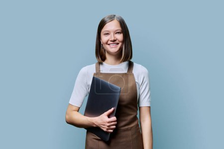 Photo for Portrait of young smiling confident woman in an apron holding laptop in hands, looking at camera on blue studio background. Worker, startup, small business, job, service sector, staff, youth concept - Royalty Free Image