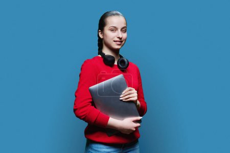 Photo for Portrait of teenage girl student in headphones with laptop in hands on blue studio background. Smiling female teenager in red looking at camera. Adolescence, education leisure lifestyle technology - Royalty Free Image
