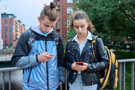 Photo for Teen friends guy and girl standing together holding smartphones looking at screen using mobile phones outdoor on city. Internet digital technology applications for leisure study communication - Royalty Free Image