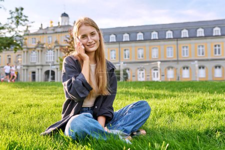 Photo for Young smiling cute female with smartphone sitting on grass, lawn in sunset light, copy space. Teenage attractive relaxed girl student talking on phone, looking at camera - Royalty Free Image