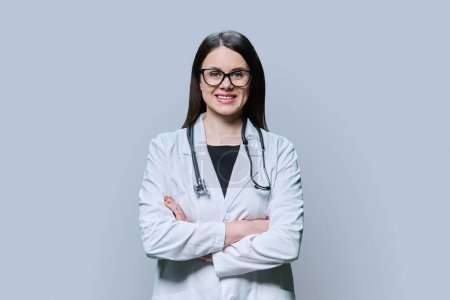Téléchargez les photos : Portrait d'une femme médecin amicale et confiante en manteau blanc sur fond de studio gris. Médecin souriant avec bras croisés regardant la caméra. Médecine, personnel médical, traitement, concept de santé - en image libre de droit