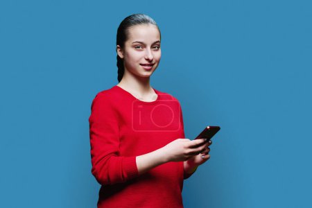 Photo for Teen girl with smartphone in hands looking at camera, on blue background. Smiling positive teenage high school student wearing red clothes. Adolescence, education leisure lifestyle technology concept - Royalty Free Image