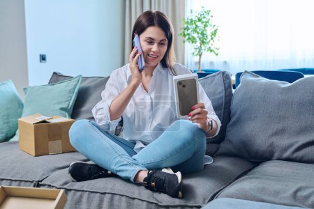 Photo for Young woman sitting on sofa at home, living room, unpacking cardboard box with online shopping, buying new mobile phone smartphone. Internet shopping, postal delivery, delivery service - Royalty Free Image