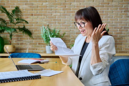 Mature business woman reading important letter, rejoicing at good news, sitting in office. Business work banking services taxes, contract statement agreement investment deal, paperwork correspondence