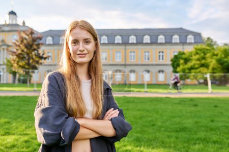 Photo for Portrait of young teenage girl student posing outdoors, background of educational building. Smiling female in glasses with textbooks standing on grass on lawn. Youth, education, college, university - Royalty Free Image