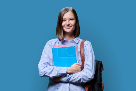 Téléchargez les photos : Portrait d'une étudiante souriante regardant une caméra sur fond de studio bleu. Une vingtaine de femmes à la mode avec sac à dos et manuels scolaires. Éducation, jeunesse, université collégiale concept - en image libre de droit