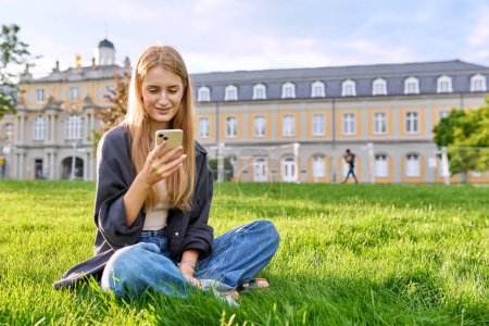 Photo for Young smiling cute female with smartphone sitting on grass, lawn in sunset light, copy space. Teenage relaxed girl student using phone, technology mobile applications for study leisure communication - Royalty Free Image