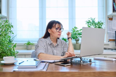 Photo for Middle-aged serious woman working at computer laptop in home office. Mature female sitting at desk typing and looking at monitor. Remote business, teaching, work, blogging, freelance - Royalty Free Image