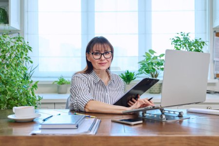 Foto de Retrato de una mujer madura que trabaja en casa en un ordenador portátil. Mujer sonriente de mediana edad que usa anteojos mirando a la cámara mientras está sentada en el escritorio de la oficina en casa. Trabajo de enseñanza de negocios remoto, freelance - Imagen libre de derechos