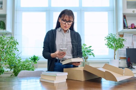 Photo for Middle aged woman with smartphone unpacking box with books, purchase from bookstore, online shopping, in home interior. Unpacking, postal delivery, online shopping, books literature concept - Royalty Free Image
