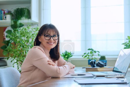 Foto de Retrato de una mujer madura que trabaja en casa en un ordenador portátil. Mujer sonriente de mediana edad que usa anteojos mirando a la cámara mientras está sentada en el escritorio de la oficina en casa. Trabajo de enseñanza de negocios remoto, freelance - Imagen libre de derechos