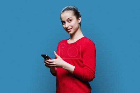 Photo for Teen girl with smartphone in hands looking at camera, on blue background. Smiling positive teenage high school student wearing red clothes. Adolescence, education leisure lifestyle technology concept - Royalty Free Image