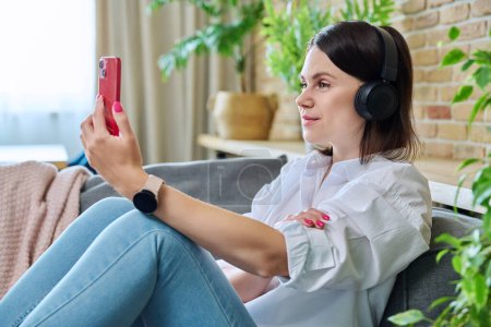 Photo for Young woman in headphones having video call chat conference, sitting on sofa at home using smartphone. Female recording video vlog blog, talking with friends, study work lifestyle - Royalty Free Image