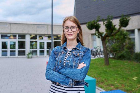 Photo for Portrait of confident teenage high school student girl with backpack, outdoor. Smiling teen schoolgirl looking at camera with crossed arms, school building background. Adolescence education knowledge - Royalty Free Image