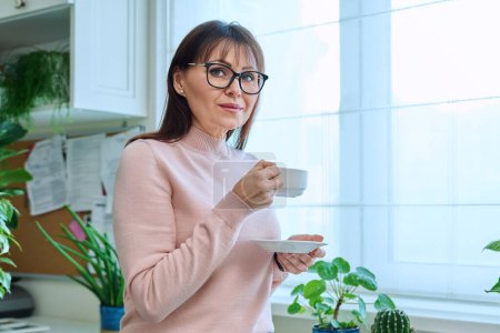 Foto de Retrato de una mujer de mediana edad sosteniendo una taza de café mirando a la cámara. Sonriente tranquila relajado hembra madura en gafas en casa cerca de la ventana. 40 años de edad, estilo de vida, concepto de salud - Imagen libre de derechos