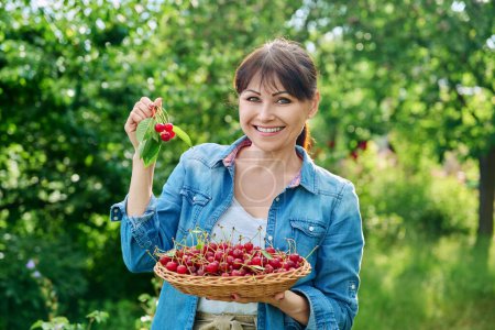 Photo for Happy woman with harvest of red ripe cherries looking at camera in summer sunny garden. Harvesting, farming, gardening, farmers market, healthy natural vitamin organic eco fruits, food concept - Royalty Free Image