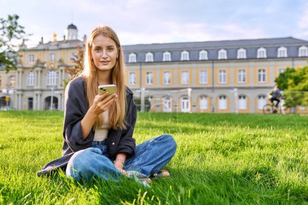 Foto de Joven sonriente linda hembra con teléfono inteligente sentado en la hierba, césped en la luz del atardecer, espacio de copia. Adolescente estudiante chica relajada utilizando el teléfono, aplicaciones móviles de tecnología para el estudio de la comunicación de ocio - Imagen libre de derechos