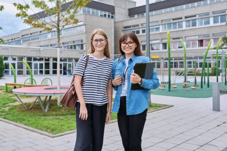 Photo for Teacher and teenage schoolgirl looking at camera together outdoor, school building background. Meeting communication student girl with backpack and mentor counselor. Education, adolescence, learning - Royalty Free Image