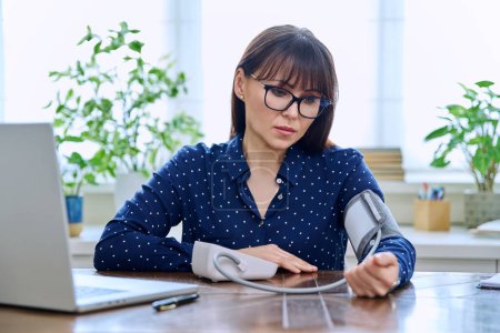 Téléchargez les photos : Femme d'âge moyen mesurant sa tension artérielle, assise au bureau avec un ordinateur sur le lieu de travail. Santé, soins, personnes matures concept - en image libre de droit