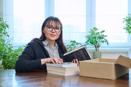 Photo for Middle-aged satisfied joyful woman with new books unpacked from cardboard box from an online store. Online shopping, unpacking, reading fiction educational non-fiction literature, hobby and leisure - Royalty Free Image