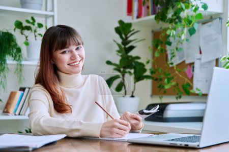 Photo for Young female college student studying at home at desk using computer laptop, writing in notebook, smiling looking at camera. E-learning, education, technology, knowledge, youth concept - Royalty Free Image