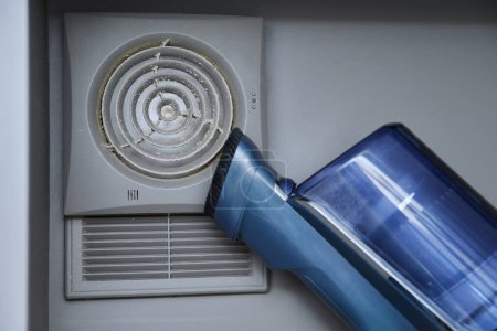 Photo for Cleaning wall-mounted dusty hood ventilation grill in bathroom with vacuum cleaner. Housekeeping, housework, housecleaning, cleaning service concept - Royalty Free Image