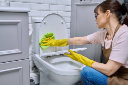 Téléchargez les photos : Femme femme au foyer femme de service de nettoyage dans des gants de protection tablier en utilisant un agent de nettoyage détergent chiffon professionnel pour nettoyer les toilettes dans la salle de bain. Désinfection hygiénique entretien ménager - en image libre de droit
