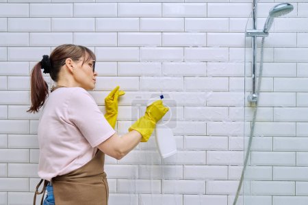 Photo for Middle-aged woman in apron, gloves cleaning bathroom with professional rag detergent spray cleans tiles in shower. Female housewife cleaning house, service worker at workplace. Housekeeping housework - Royalty Free Image