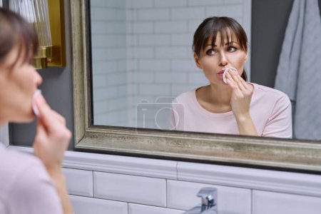 Téléchargez les photos : Femme d'âge moyen avec coton tampon nettoie son visage, lèvres de rouge à lèvres, regarder dans le miroir à la maison dans la salle de bain. Age mûr, routines beauté, concept de démaquillage - en image libre de droit