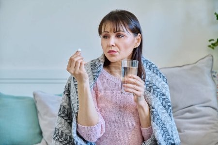 Kranke reife Frau nimmt Tablette mit Glas Wasser und sitzt zu Hause unter einer Decke. Medikamente, Gesundheit, Behandlungskonzept