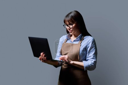 Photo for Business woman owner entrepreneur worker in an apron using laptop computer profile view on gray studio background. Mobile apps applications for business communication, job, staff management technology - Royalty Free Image