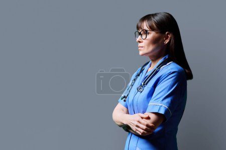 Téléchargez les photos : Portrait d'une infirmière sérieuse d'âge moyen confiante aux bras croisés regardant de profil sur fond de studio gris, espace de copie. Services médicaux, santé, assistance professionnelle, soins médicaux - en image libre de droit
