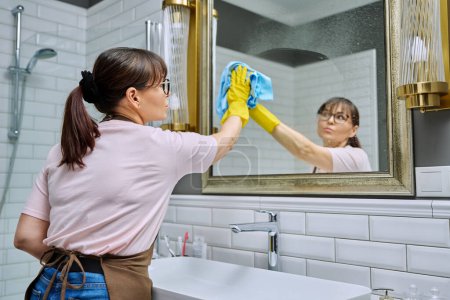 Foto de Mujer de mediana edad en un delantal, guantes de baño de limpieza, con spray detergente trapo profesional limpia el espejo. Mujer ama de casa limpieza casa, trabajador de servicios en el lugar de trabajo. Trabajo doméstico doméstico - Imagen libre de derechos