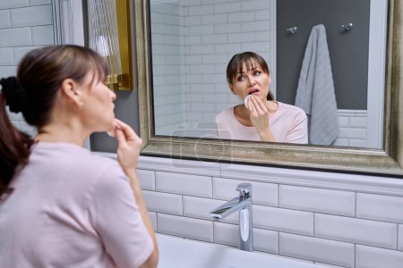 Foto de Mujer de mediana edad con almohadilla de algodón limpia su cara, labios de lápiz labial, mirando en el espejo en casa en el baño. Edad madura, rutinas de belleza, concepto de eliminación de maquillaje - Imagen libre de derechos