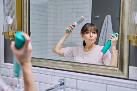 Téléchargez les photos : Femme d'âge moyen avec peigne de laque à cheveux regardant dans le miroir, faisant coiffure, dans la salle de bain. Beauté, âge, cosmétiques, accessoires capillaires - en image libre de droit