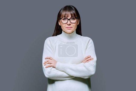 Photo for Middle-aged beautiful serious woman in white sweater on gray studio background. Confident mature female in glasses with crossed arms looking at camera. Beauty, lifestyle, business, work, 40s people - Royalty Free Image