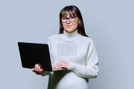 Téléchargez les photos : Belle femme d'âge moyen tenant un ordinateur portable regardant la caméra sur fond de studio gris. Sourire femme heureuse en pull blanc à l'aide d'un ordinateur. Technologies pour les entreprises communications de travail loisirs - en image libre de droit
