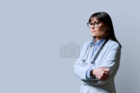 Photo for Serious mature confident woman doctor in white lab coat with stethoscope, profile view, gray background, copy space. Healthcare, medicine, staff, treatment, medical services concept - Royalty Free Image