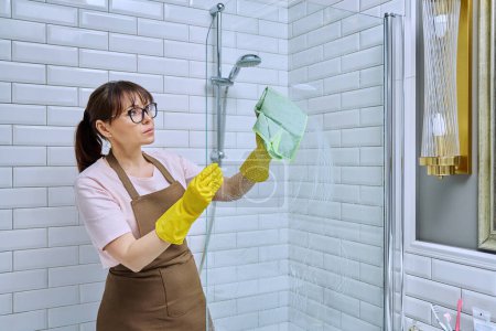 Photo for Middle-aged woman in apron, gloves cleaning bathroom with professional rag detergent spray cleans glass in shower. Female housewife cleaning house, service worker at workplace. Housekeeping housework - Royalty Free Image