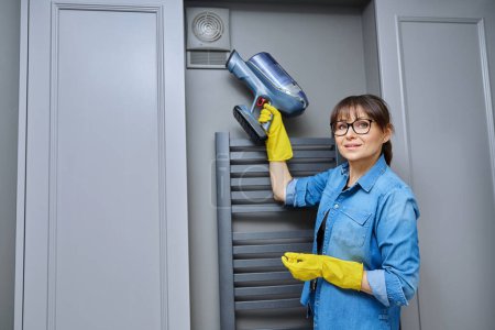 Photo for Woman cleaning wall-mounted dusty hood ventilation grill in bathroom with vacuum cleaner. Housekeeping, housework, housecleaning, cleaning service concept - Royalty Free Image