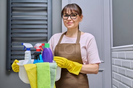 Foto de Mujer de mediana edad en delantal de guantes con lavabo de detergentes en el baño. Mujer preparándose para la limpieza rutinaria de la casa, empleada de servicio de limpieza posando en el lugar de trabajo. Limpieza de las tareas domésticas - Imagen libre de derechos