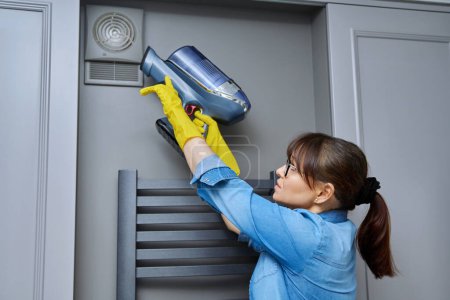 Woman cleaning wall-mounted dusty hood ventilation grill in bathroom with vacuum cleaner. Housekeeping, housework, housecleaning, cleaning service concept