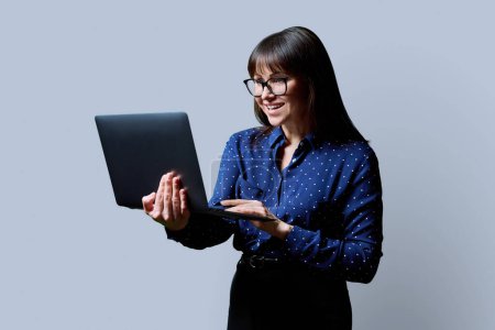 Foto de Sonriente mujer de mediana edad de negocios utilizando el ordenador portátil en fondo de estudio gris. Confiada exitosa madura sosteniendo la computadora en las manos. Negocios, trabajo, trabajo, estilo de vida, tecnología, concepto de personas de 40 años - Imagen libre de derechos