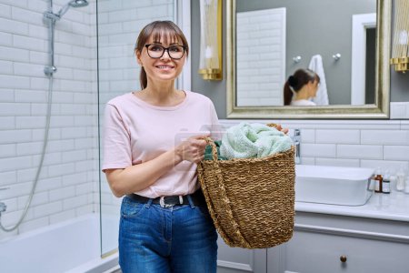 Téléchargez les photos : Femme d'âge moyen avec panier de serviettes pour se laver à la maison dans la salle de bain, souriante femme regardant la caméra. Entretien ménager, blanchisserie, vie domestique, concept de style de vie - en image libre de droit