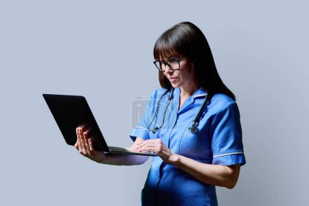 Photo for Serious female nurse using laptop computer, profile view on gray studio background. Mobile apps applications technologies in medical services, health, professional assistance, medical care concept - Royalty Free Image