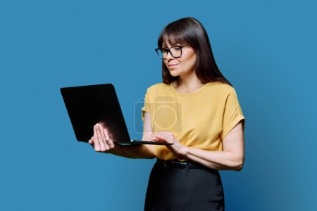 Photo for Middle-aged business confident woman using laptop on blue background. Mature serious female teacher mentor manager worker employee director. Internet online technologies, work, electronic teaching - Royalty Free Image