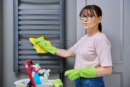 Photo for Woman doing house cleaning in bathroom, cleaning dust from heated towel rail. Housekeeping, housework, housecleaning, cleaning service concept - Royalty Free Image