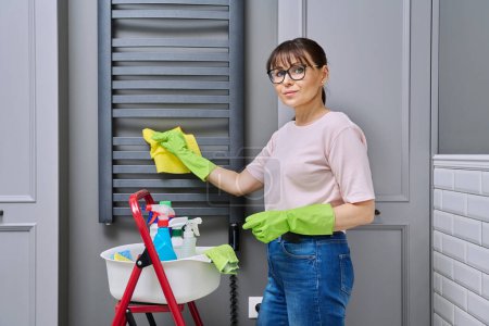 Téléchargez les photos : Femme faisant le nettoyage de la maison dans la salle de bain, nettoyer la poussière du porte-serviettes chauffant. Entretien ménager, travaux ménagers, ménage, concept de service de nettoyage - en image libre de droit