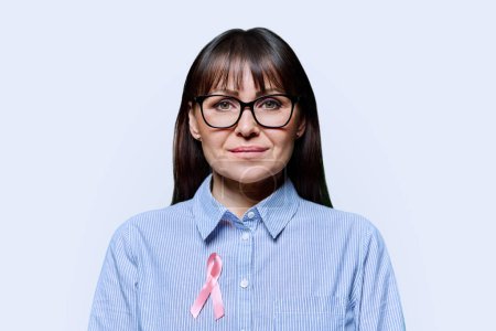 Photo for Middle-aged woman with pink ribbon on her chest symbolizing International Breast Cancer Day, on white studio background. Solidarity, patient support, health care, treatment, medicine, world cancer day - Royalty Free Image